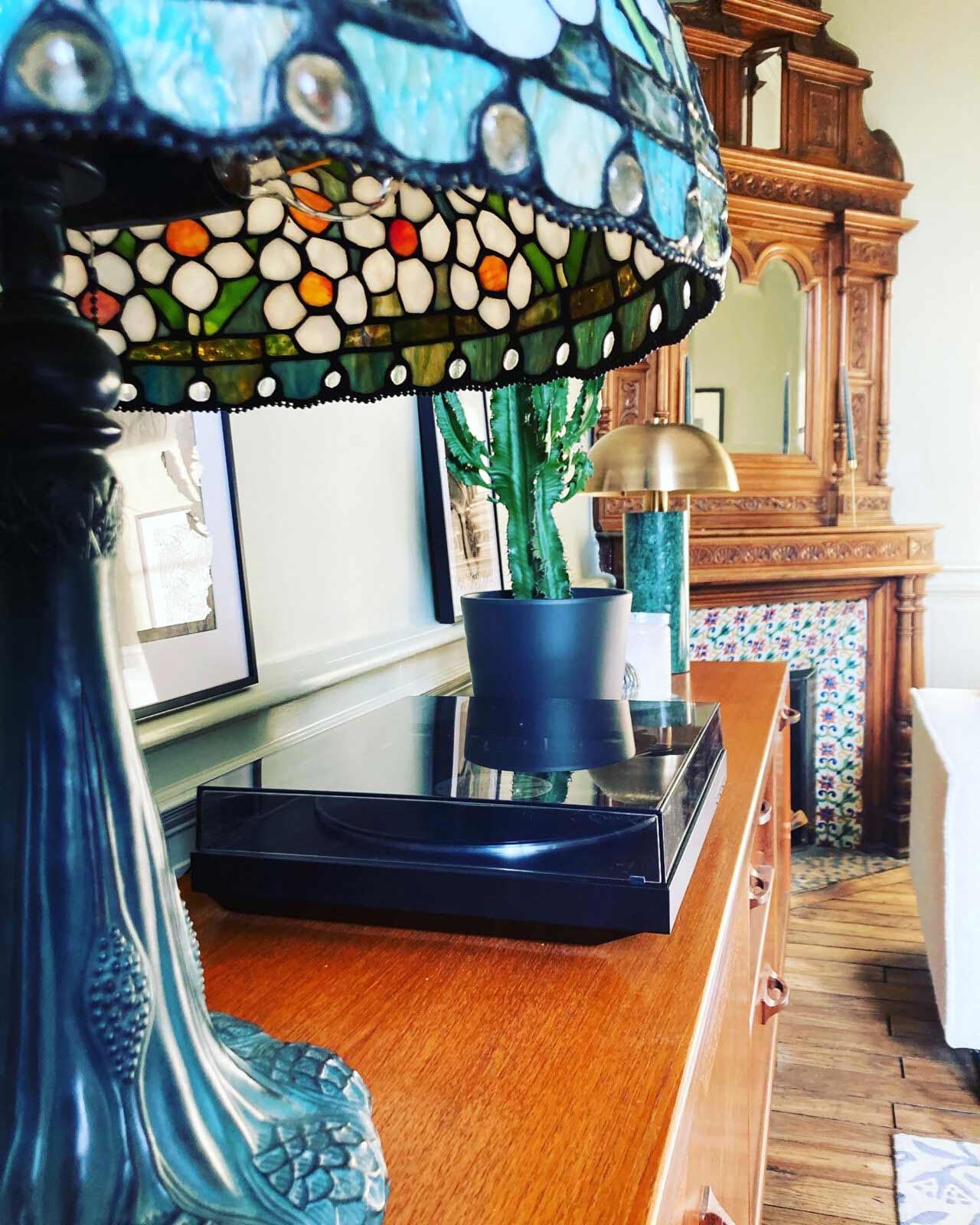 View of the decor on the top of the chest of drawers. A record player, cactus and a stain glass lamp is situated on top.